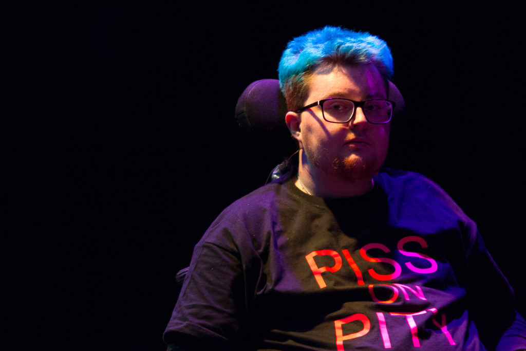 A person is seated. A wheelchair headrest is visible behind them. The image is dark, but their hair is a bright blue. Their t-shirt is blace, and says "PISS ON PITY" in bright pink. They are wearing a microphone and glasses.