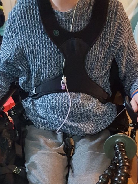 The image shows my upper body in a blue jumper, with a 4 point black harness over me, securing me at the shoulders and hips. My feeding tube is dangling.
