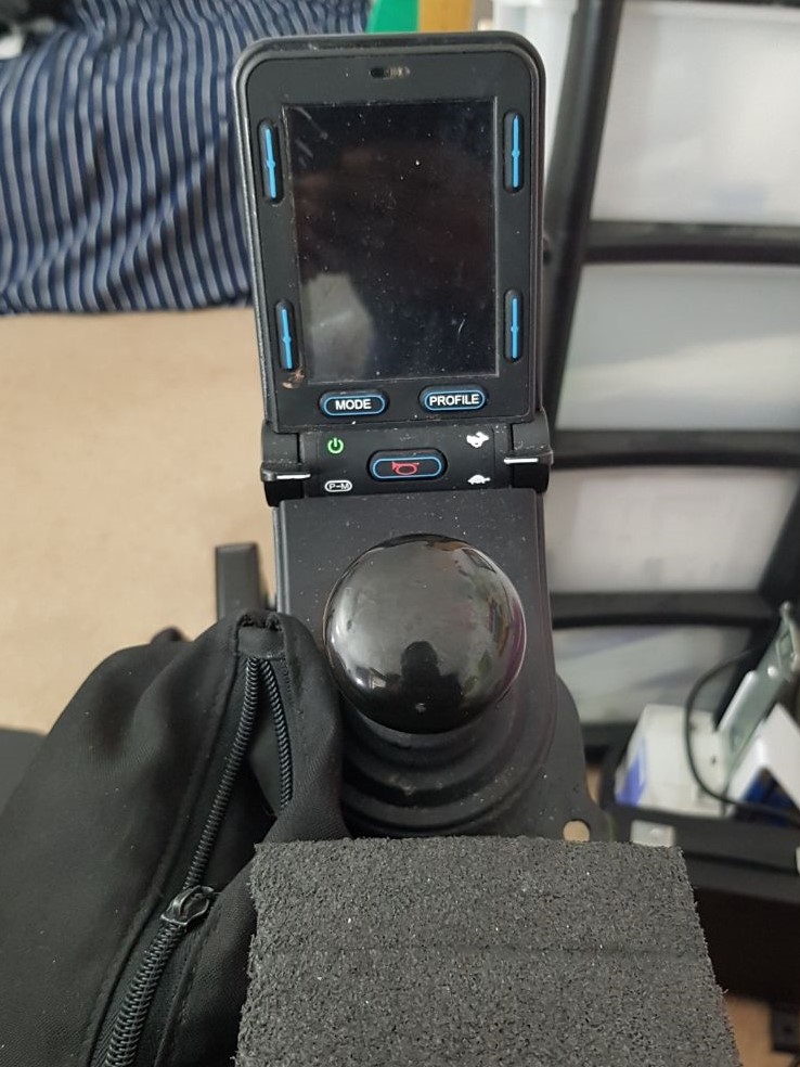 The centre of the image is a mushroom shaped joystick. Above it is a black screen, switched off. Below the wheelchair joystick is some black foam and a black bag.