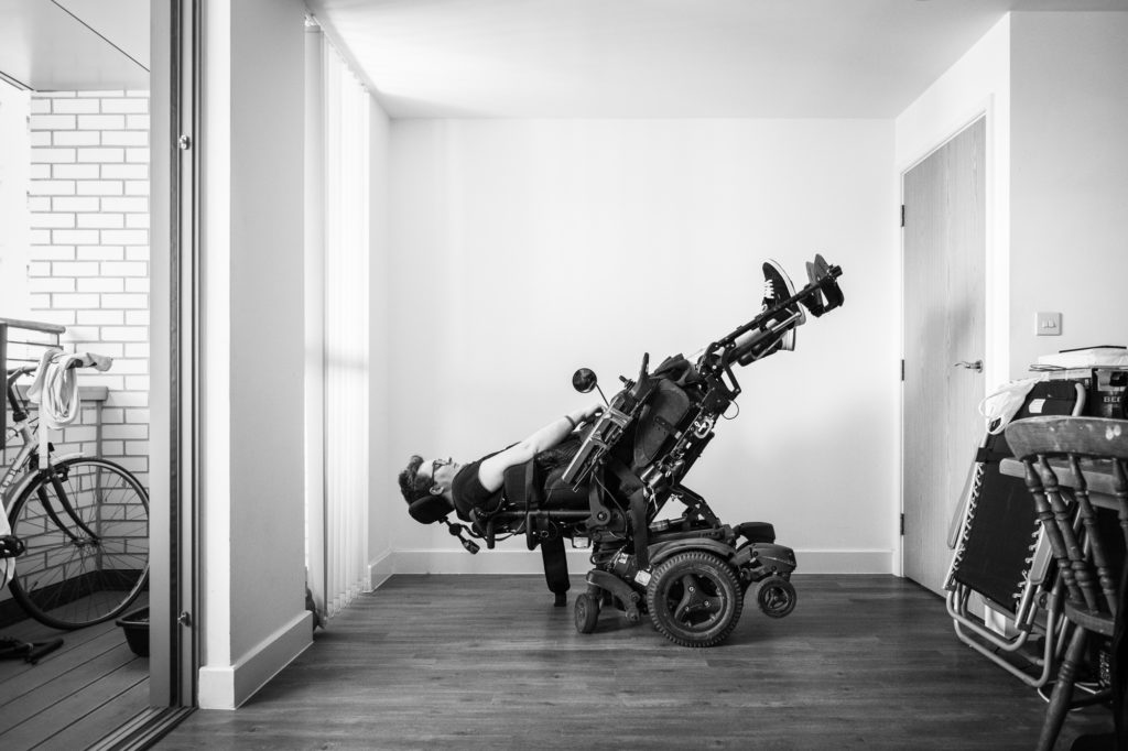 The image shows a white room. On the far left is a balcony with a bicycle on it. On the far right is some stuff. In the centre of the image, which is black and white, is a white person in an electric wheelchair. They are almost upside down, with their head near the floor and their legs extended straight upwards into the air