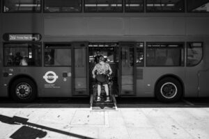 A black and white image showing a London double-decker bus. The ramp is out, and a white person in an electric wheelchair is driving down the ramp forwards