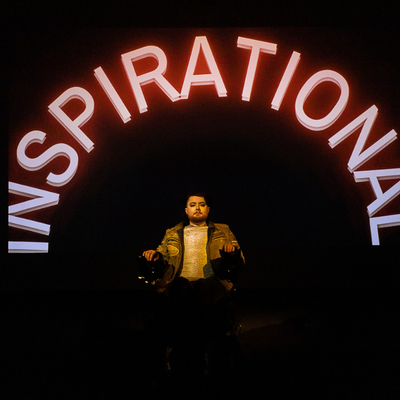 A black background with Jamie seated in their electric wheelchair, lit from the torso upwards. Above them in pink neon is the word "inspirational"