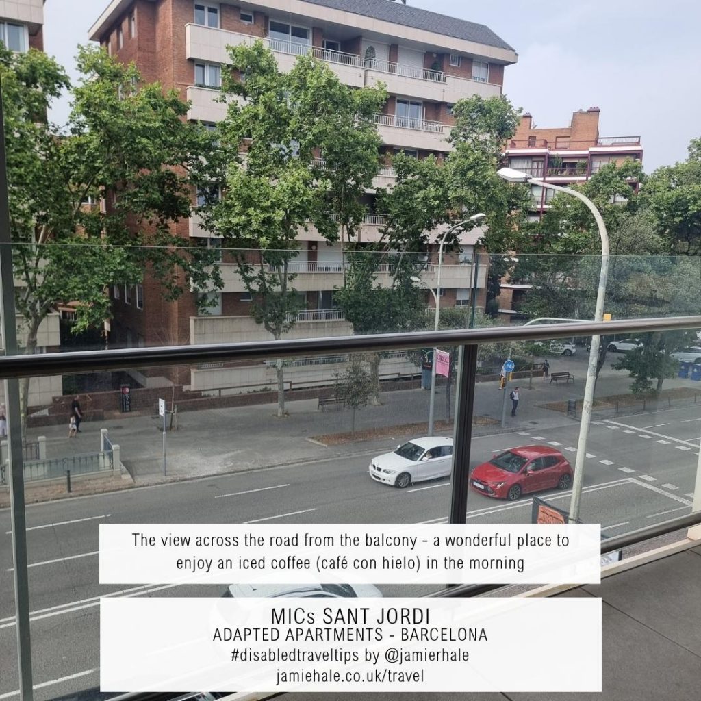 Superimposed on top of a photo of the view from a glass wall balcony across a tree line road to buildings on the other side is the text "MICs Sant Jordi, adapted apartments, Barcelona, They give us reload from the balcony, a wonderful place to enjoy an iced coffee (cafe con hielo) in the morning, #DisabledTravelTips by @jamierhale jamiehale.co.uk/travel"