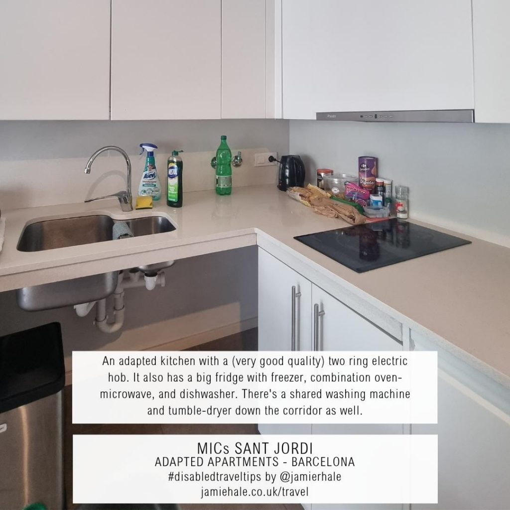 Superimposed on top of a photo of a kitchen with lots of white cupboards, white surfaces, a sink with empty space underneath it instead of a cupboard, and a hob with two rings is the text "MICs Sant Jordi, adapted apartments, Barcelona, unadapted kitchen with a very good quality to ring electric hub. It also has a big fridge with freezer, combination oven microwave, and dishwasher. There's a shared washing machine and tumble dryer down the corridor as well, #DisabledTravelTips by @jamierhale jamiehale.co.uk/travel"