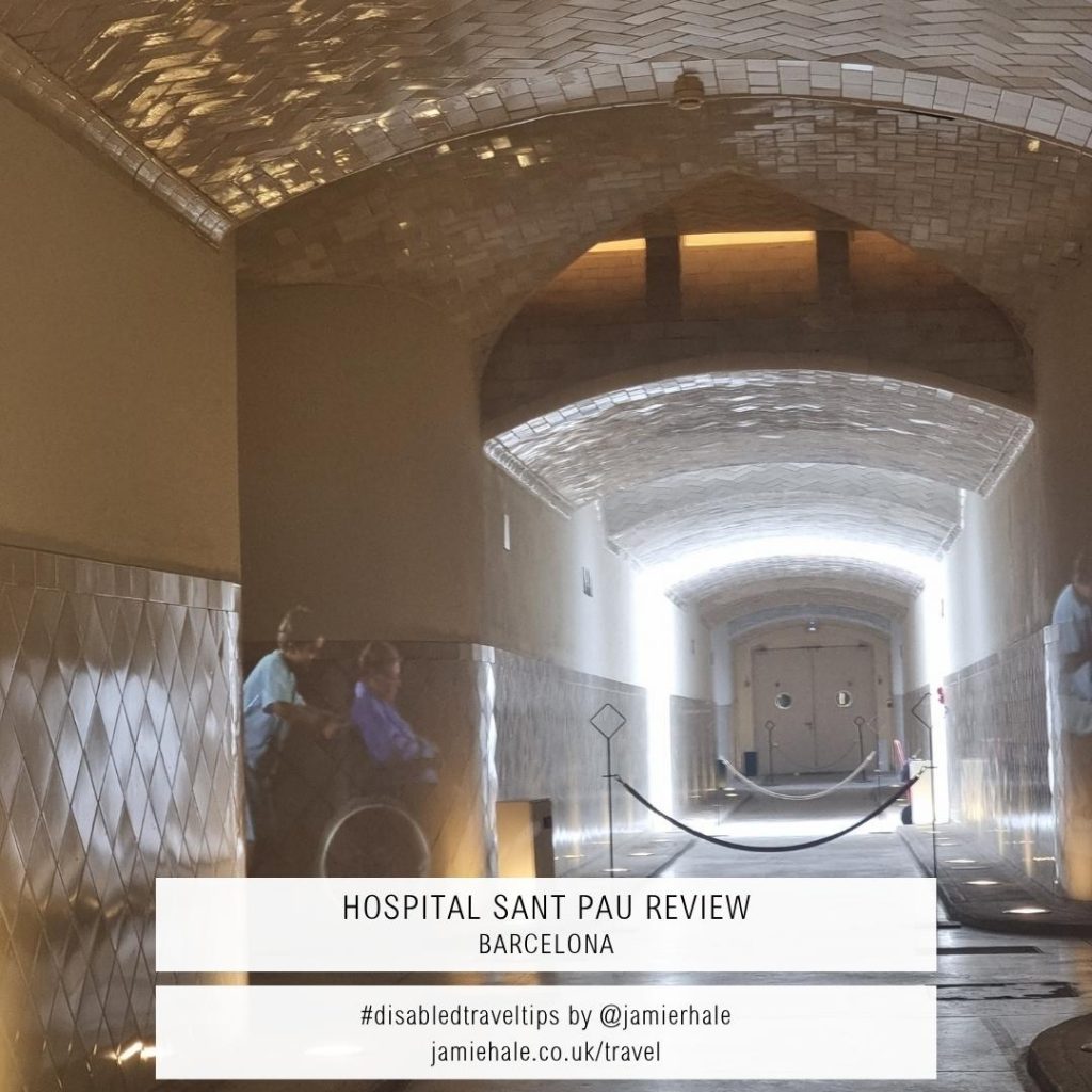 A photo taken inside a corridor, with shiny tiles along the lower half of the walls, and all over the ceiling, which are reflecting the light. There is an image projected onto the tiles of someone in a wheelchair and someone in medical clothing pushing the chair. Superimposed over the top of the photo is text reading 'Hospital Sant Pau Review Barcelona' '#disabledtraveltips by @jamierhale jamiehale.co.uk/travel'