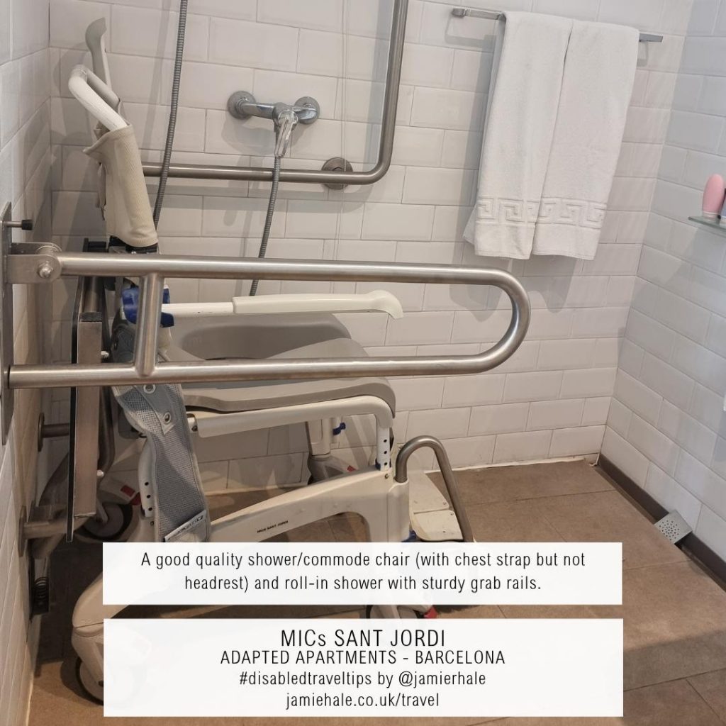 Superimposed on top of a wheeled shower chair with a high backrest and armrests, heart in a shower with a drop-down grab rail on one side of it and the wall on the other. With a towel hanging beside it. is the text "MICs Sant Jordi, adapted apartments, Barcelona, a good quality shower and commode chair with chest strap but not headrest, and roll in shower with sturdy grab rails, #DisabledTravelTips by @jamierhale jamiehale.co.uk/travel"