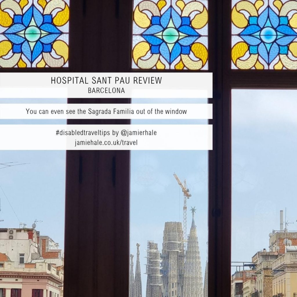 A photo taken from inside out of a window, showing the Sagrada Familia and a few other buildings. The top of the windows are blue and yellow floral stained glass panes. Superimposed over the image is text reading 'Hospital Sant Pau Review Barcelona', 'You can even see the Sagrada Familia out of the window', '#disabledtraveltips by @jamierhale jamiehale.co.uk/travel'