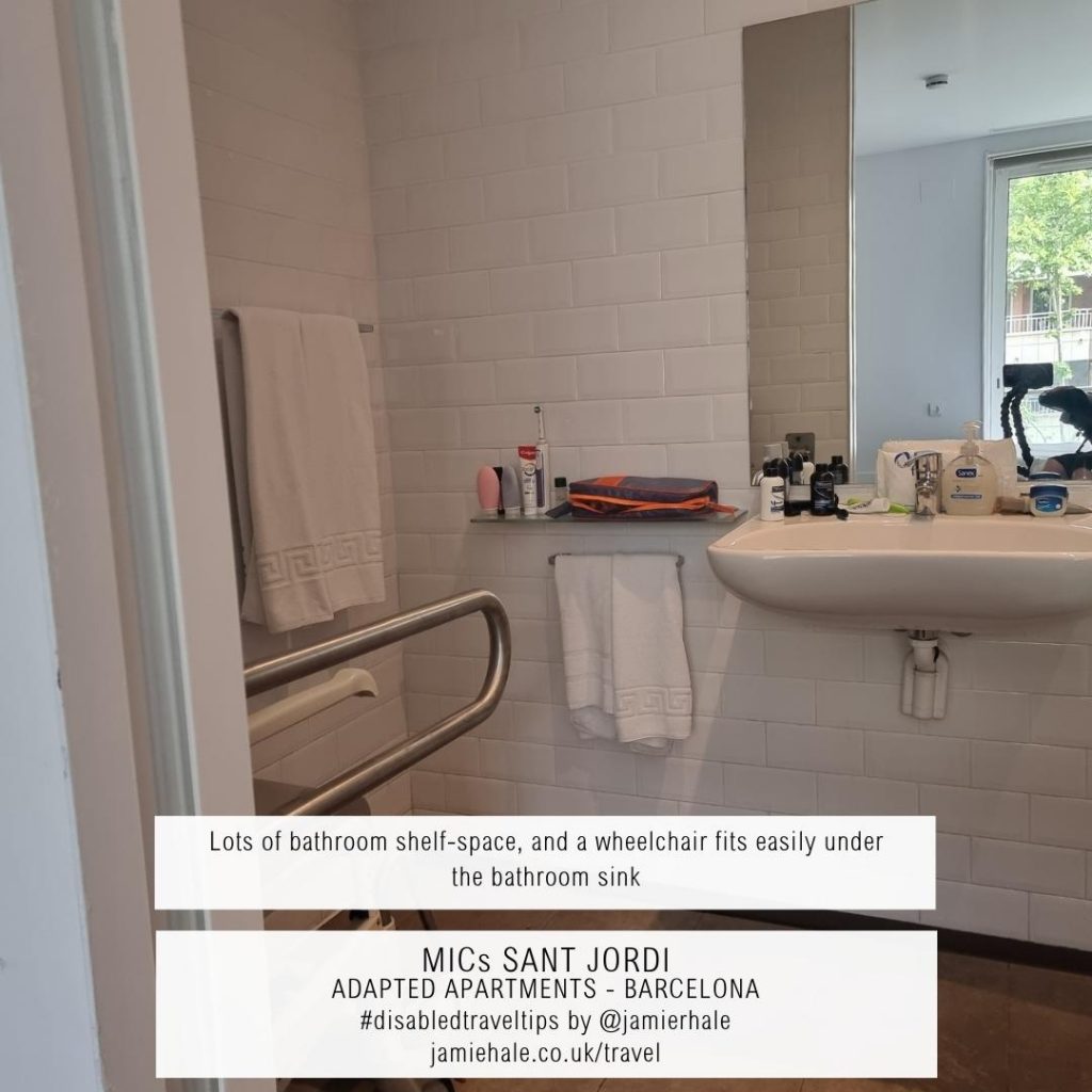 Superimposed on top of a photo of a bathroom showing a drop-down grab rail, a shelf and sink with various things on the side, lots of space under the sink, and a mirror is the text "MICs Sant Jordi, adapted apartments, Barcelona, lots of bathroom shelf space, and a wheelchair fits easily under the bathroom sink, #DisabledTravelTips by @jamierhale jamiehale.co.uk/travel"