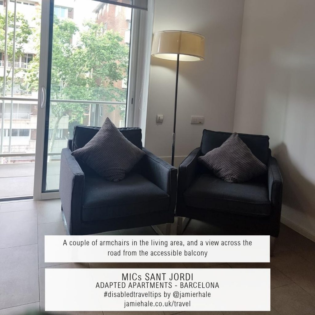 Superimposed on top of a picture of two grey armchairs, a tall lamp, and, behind them, sliding glass doors leading to a balcony is the text "MICs Sant Jordi, adapted apartments, Barcelona, a couple of armchairs in the living area, and a view across the road from the accessible balcony, #DisabledTravelTips by @jamierhale jamiehale.co.uk/travel"