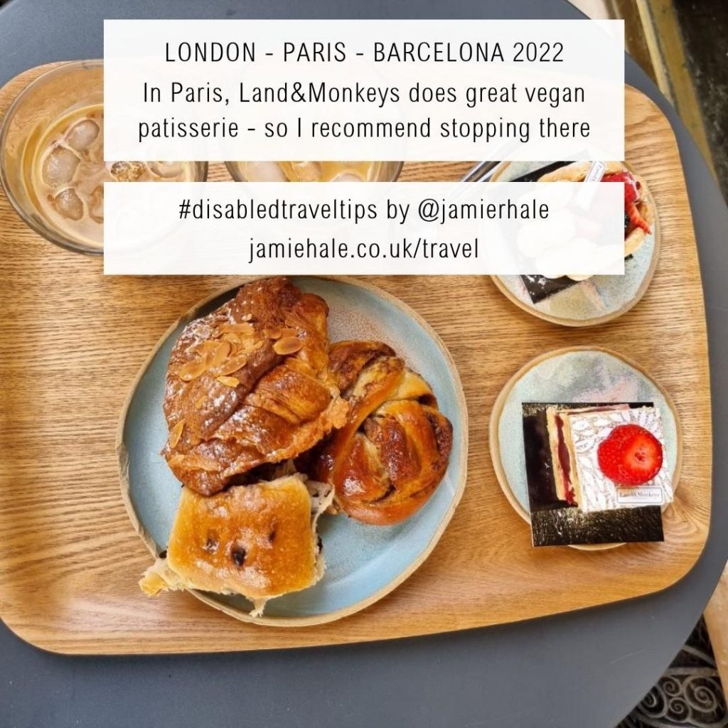 Superimposed on top of a picture of a wooden breakfast tray with a plate full of pastries, two smaller plares with layered sponge cakes, and two iced coffees is the text "LONDON-PARIS-BARCELONA 2022 In paris, Land&Monkeys does great vegan patisserie - so I recommend stopping there. #DisabledTravelTips by @jamierhale jamiehale.co.uk/travel"