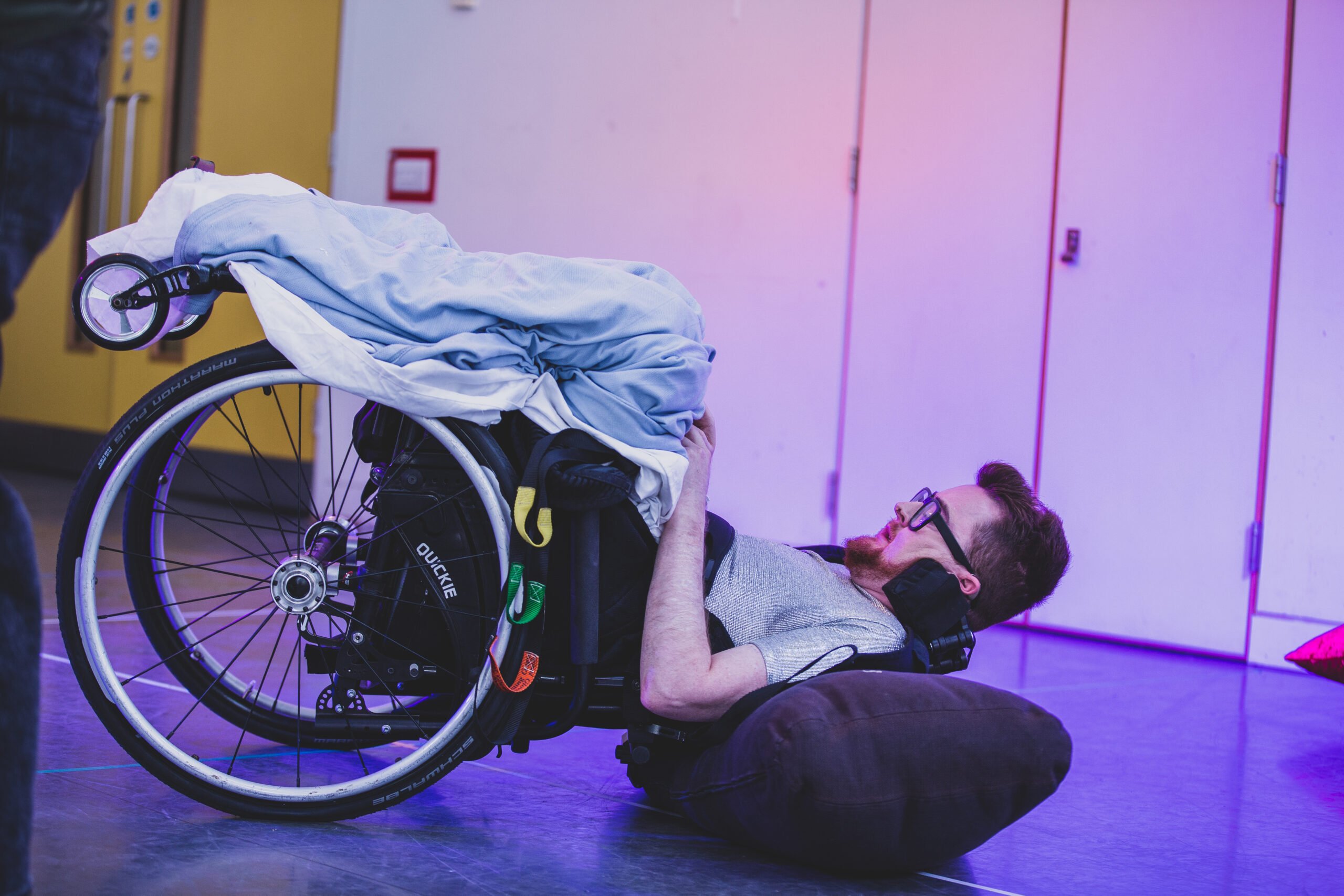 Bathed in purple light, Jamie Hale lies on their back on the floor in a manual wheelchair, a hospital blanket over their legs, to illustrate their workshops and performances