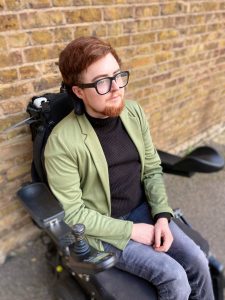 In one of their Jerwood photos, Jamie sits against a brick wall. They are in a black electric wheelchair, wearing a black rollneck and green blazer. Their ginger hair swoops across their forehead and matches their beard.