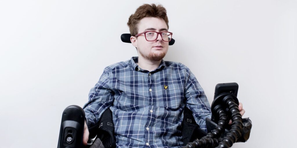 Jamie, a white person with dark red hair and a red beard in an electric wheelchair. They are wearing red glasses and a blue check shirt, and are against a white background.