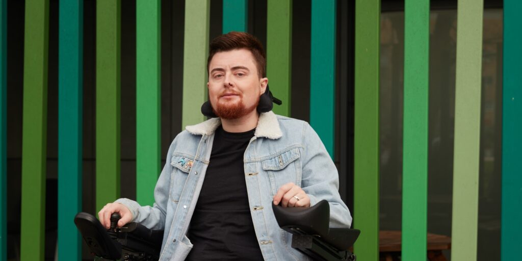 Jamie Hale, a white person with dark red hair and a red beard, in an electric wheelchair. They are wearing a blue denim jacket, and against an outdoor green and black striped background.