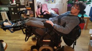 Me, stretched out in my wheelchair. My feet are on the leg-rests on the left of the image an d my head is towards the top at the right. You can see me being strapped in using a 4 point harness, with my arms resting on channel armrests.
