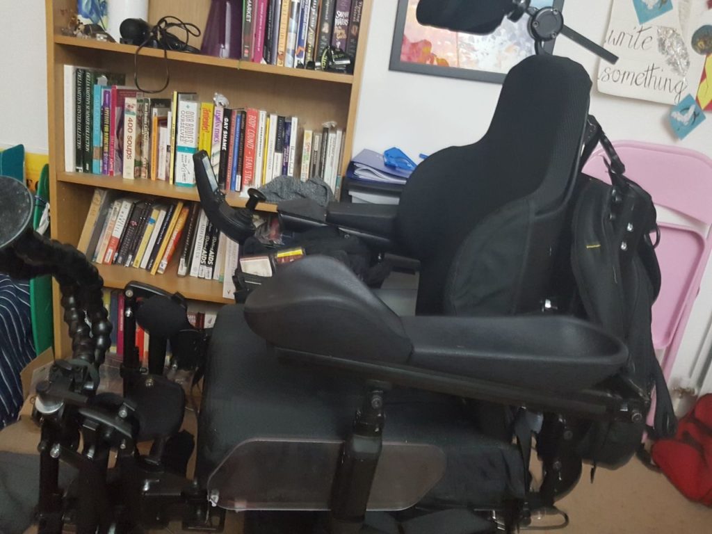 The electric wheelchair parked in front of a bookshelf, with the backrest completely upright