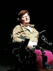 A white person with dark red hair sits in a black electric wheelchair against a black background. They are wearing pink jeans and a leopard print jacket
