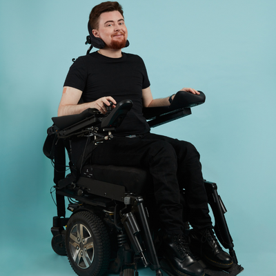 A full-body shot of Jamie in their electric wheelchair against a blue background