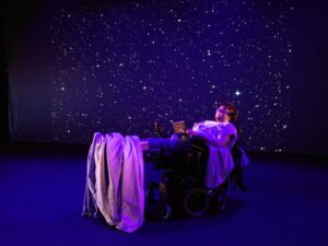 The picture has a dark background with white pinpricks that are stars. Jamie is lying in a hospital bed, bathed in piunk and blue light.