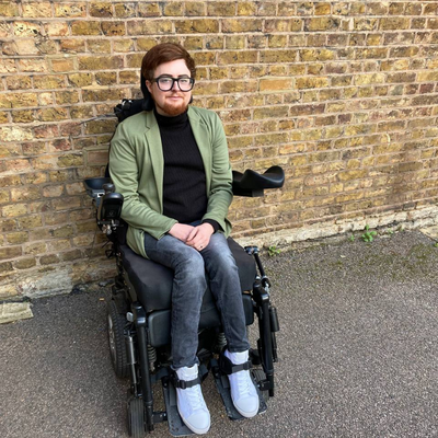 A brick wall behind Jamie, a white person in an electric wheelchair, wearing a khaki blazer and black skinny jeans with white trainers