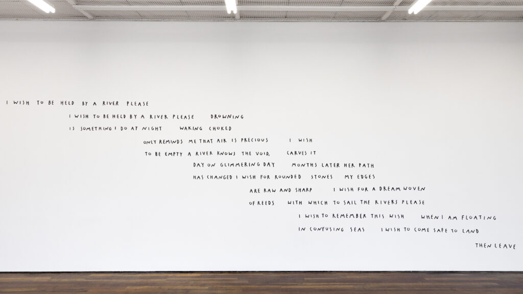 A white wall with text "I wish to be held by a river please i wish to be held by a river please drowning is something i do at night waking choked only reminds me that air is precious i wish to be empty a river knows the void, carves it day on glimmering day months later her path has changed i wish for rounded stones my edges are raw and sharp i wish for this not to end for me i wish to be free i wish for a dream woven of reeds with which to sail the rivers please i wish to remember this wish when i am floating in confusing seas i wish to come safe to land then leave i wish to be floating forever please"