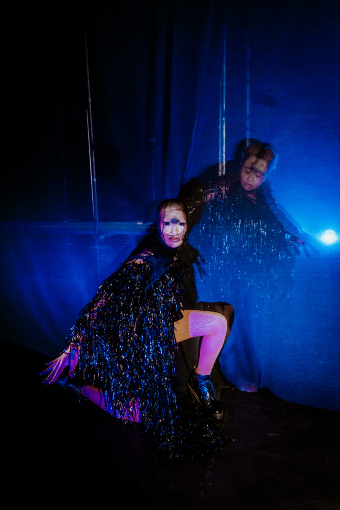 Ila in front of a curtain and Ray behind it, a dark blue gauziness over the image at Transpose Subverse in the Pit at the Barbican in 2025