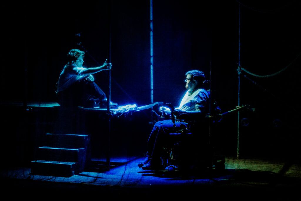 Jamie Hale and CN Lester look at each other, CN on scaffolding, Jamie in their chair, in deep blue light