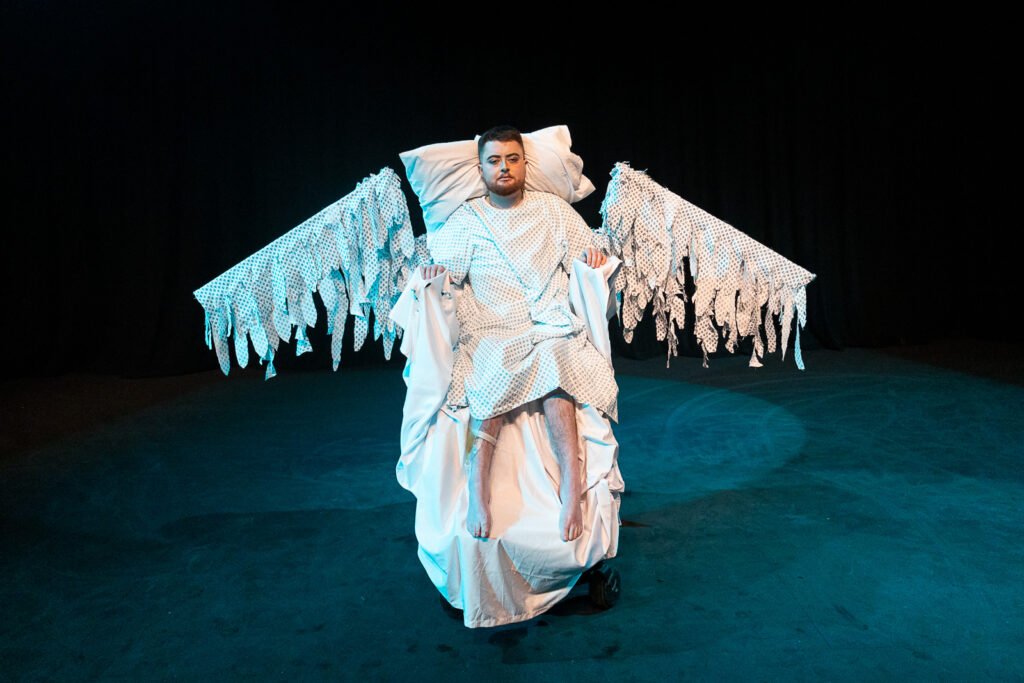 Jamie Hale, a white electric wheelchair user lies on a white surface, sat up. They are wearing a hospital gown and have wings outstretched to either side of their shoulders, also in hospital gown material!