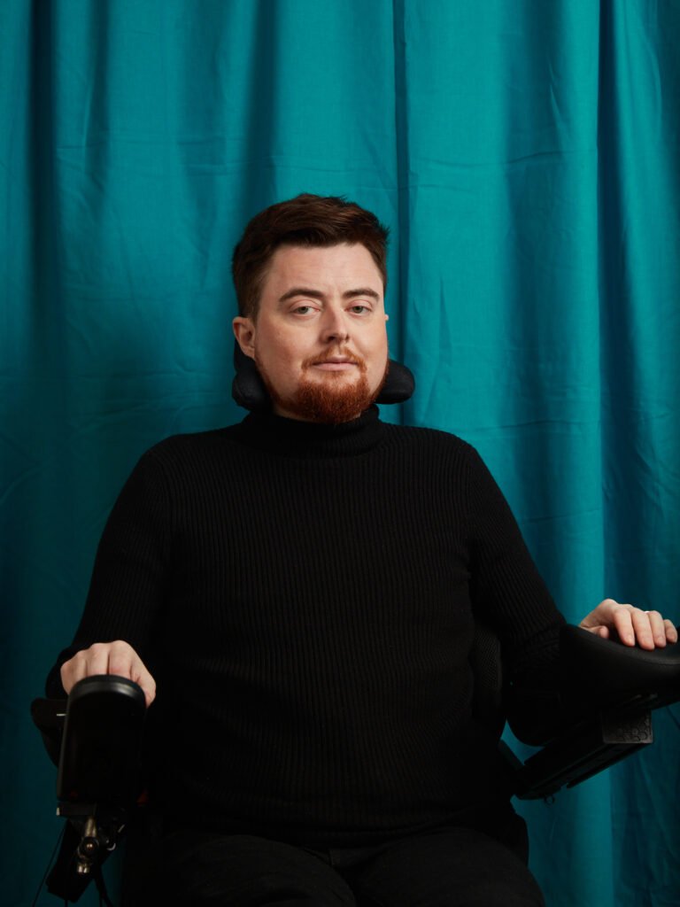 Jamie Hale, a white electric wheelchair user wearing all black sits in front of a rich turquoise curtain.

