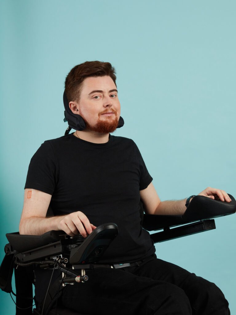 Jamie Hale, a white electric wheelchair user with dark red hair, in front of a blue background.