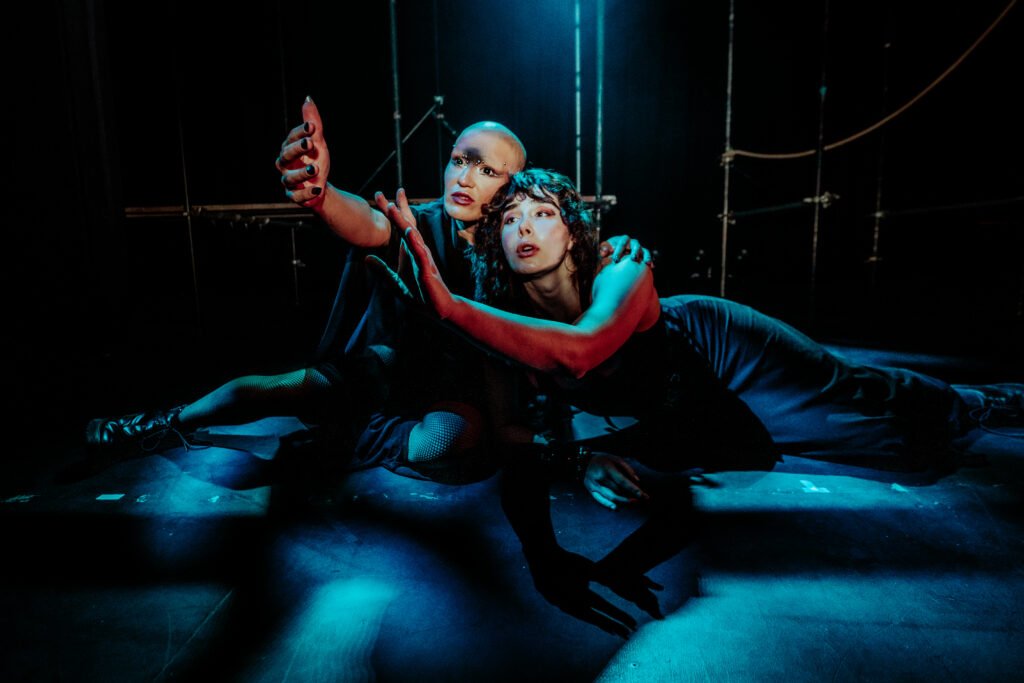 ILĀ and Coda Nicolaeff sit on the floor, leaning and reaching forward with both arms outstretched. They lean into each other, both wearing black.
